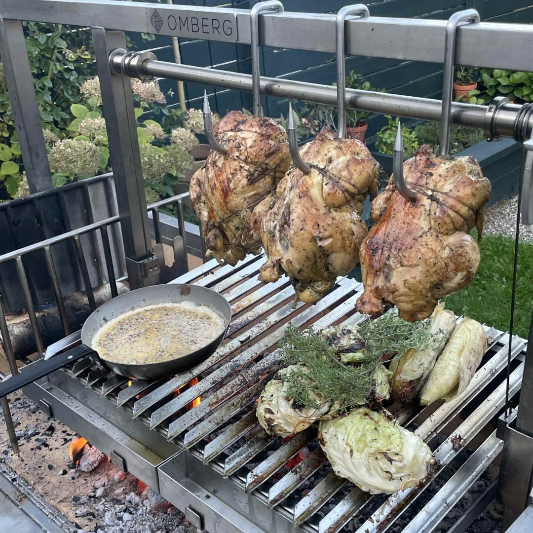 Hele kip wordt samen met maïs geroosterd, salade hangt aan vleeshaken boven de Omberg Asado 1200 Parilla .
