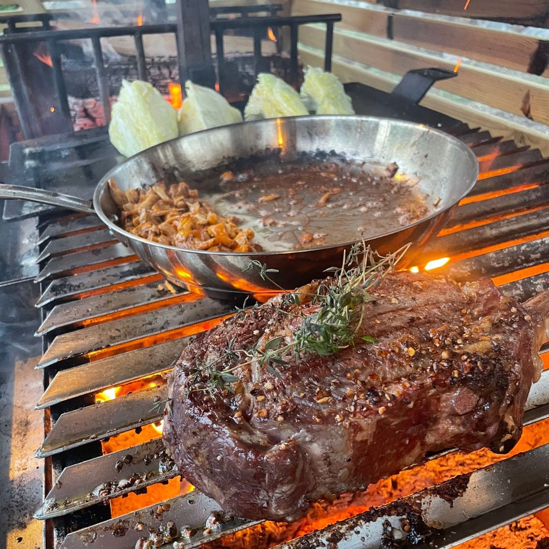 Ribeye-Steak mit Knochen und Pilze, gebraten in einer Pfanne über glühender Holzkohle auf Omberg Asado Parrilla-Grill
